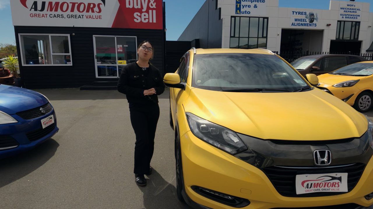 A presenter standing beside a yellow Honda on the AJ Motors car yard in Woolston